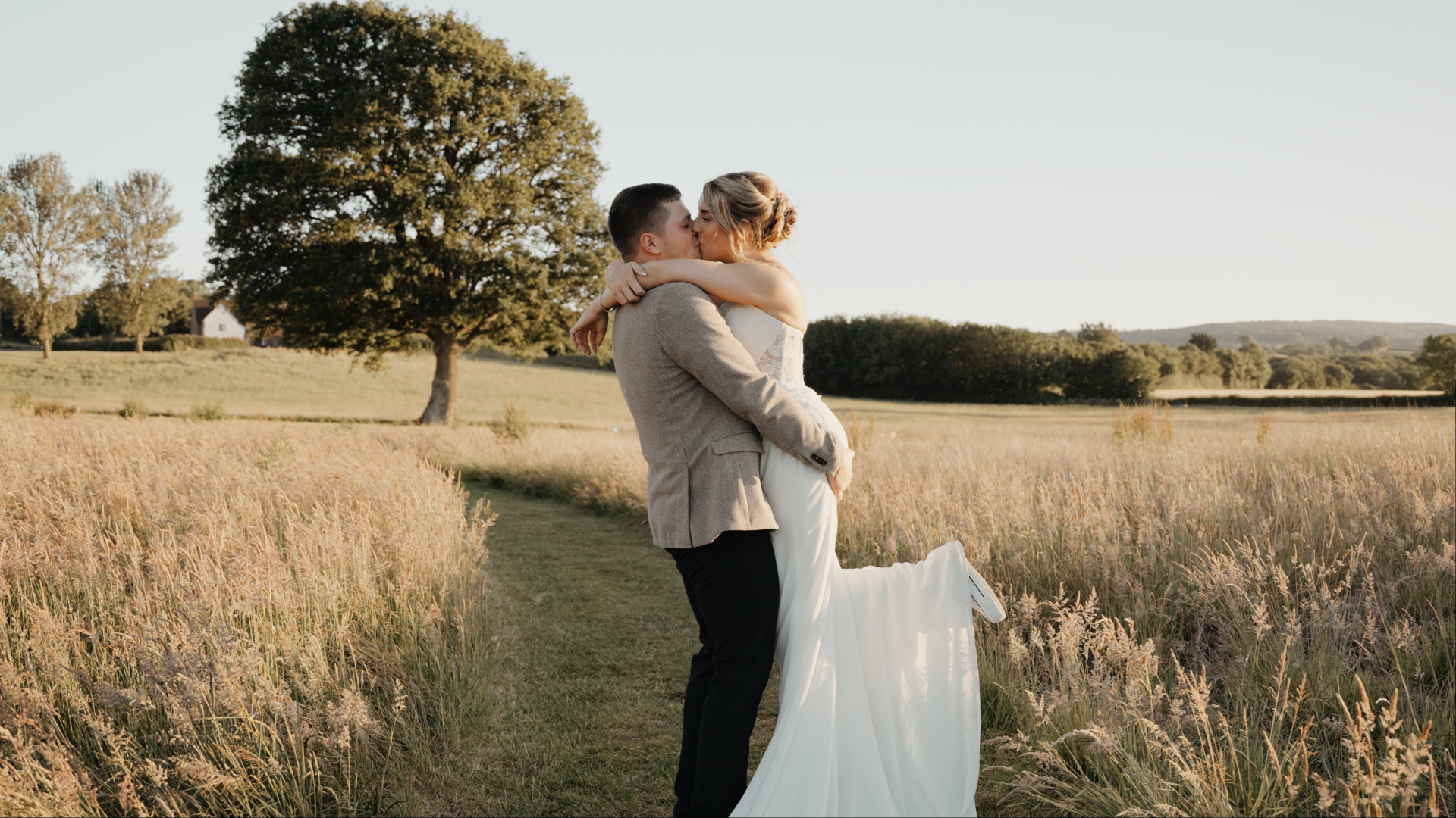 A couple embracing in a field at sunset, with the groom lifting the bride, capturing a perfect wedding videography moment. The bride wears a white dress and the groom a light brown jacket. A large tree and distant house add to the serene romance of the scene.