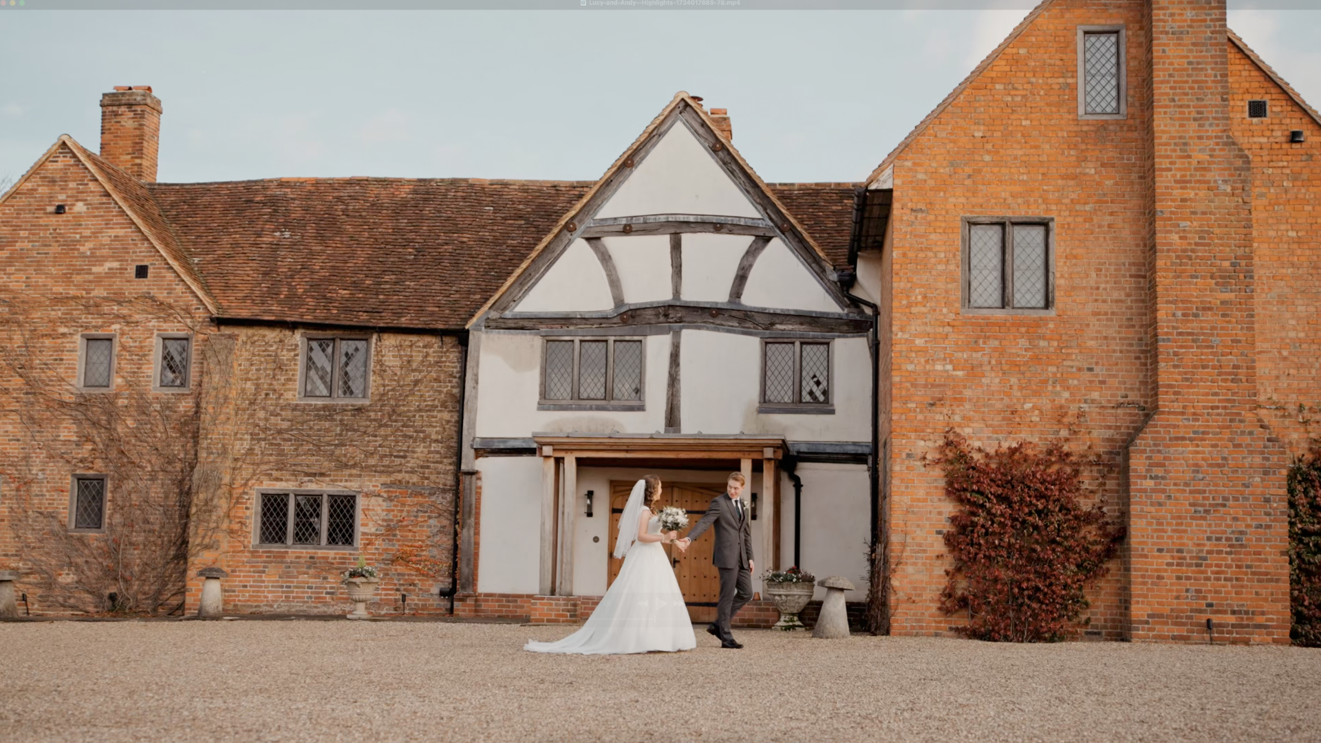 A bride and groom stand hand in hand before the majestic Lillibrooke Manor, its historic brick and timber architecture adding charm to their serene, elegant moment. The brides white gown and the grooms suit enhance this picturesque scene.