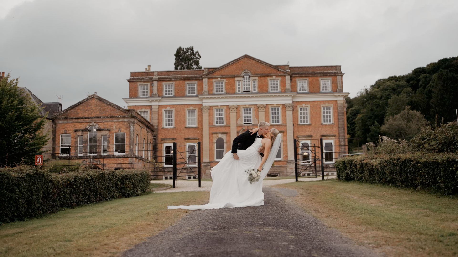 The bride and groom are embracing dramatically on the path before the grandeur of Crowcombe Court, a historic brick mansion. With a cloudy sky above, she holds a bouquet in her white gown, while he stands by her side in a black suit.