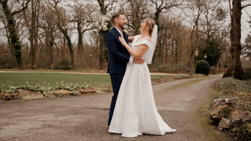 A bride and groom stand on a path at Berwick Lodge, surrounded by trees. The bride, in a white dress and veil, holds a bouquet while the groom, clad in a navy suit, smiles back at her. The lush greenery frames their serene moment perfectly.