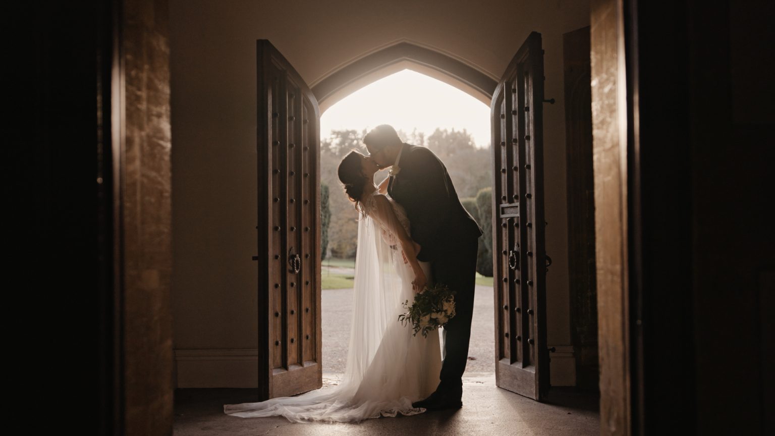 A bride and groom kiss in silhouette at St Audries Park, framed by large open wooden doors with intricate designs. The bride wears a long veil and holds a bouquet. Sunlight filters through the opening, creating a romantic, warm glow around the couple.