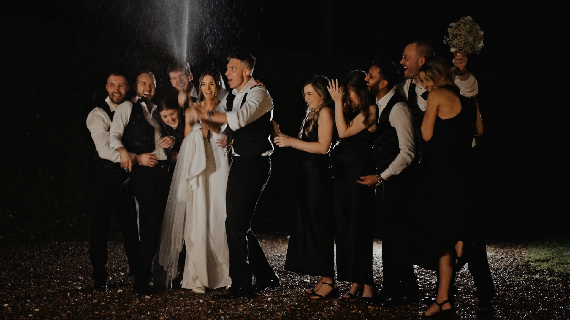 A joyful wedding party celebrates at night as the groom pops a champagne bottle, with the bride and guests laughing and reacting to the spray. Everyone is dressed in formal attire, creating a festive and energetic atmosphere.