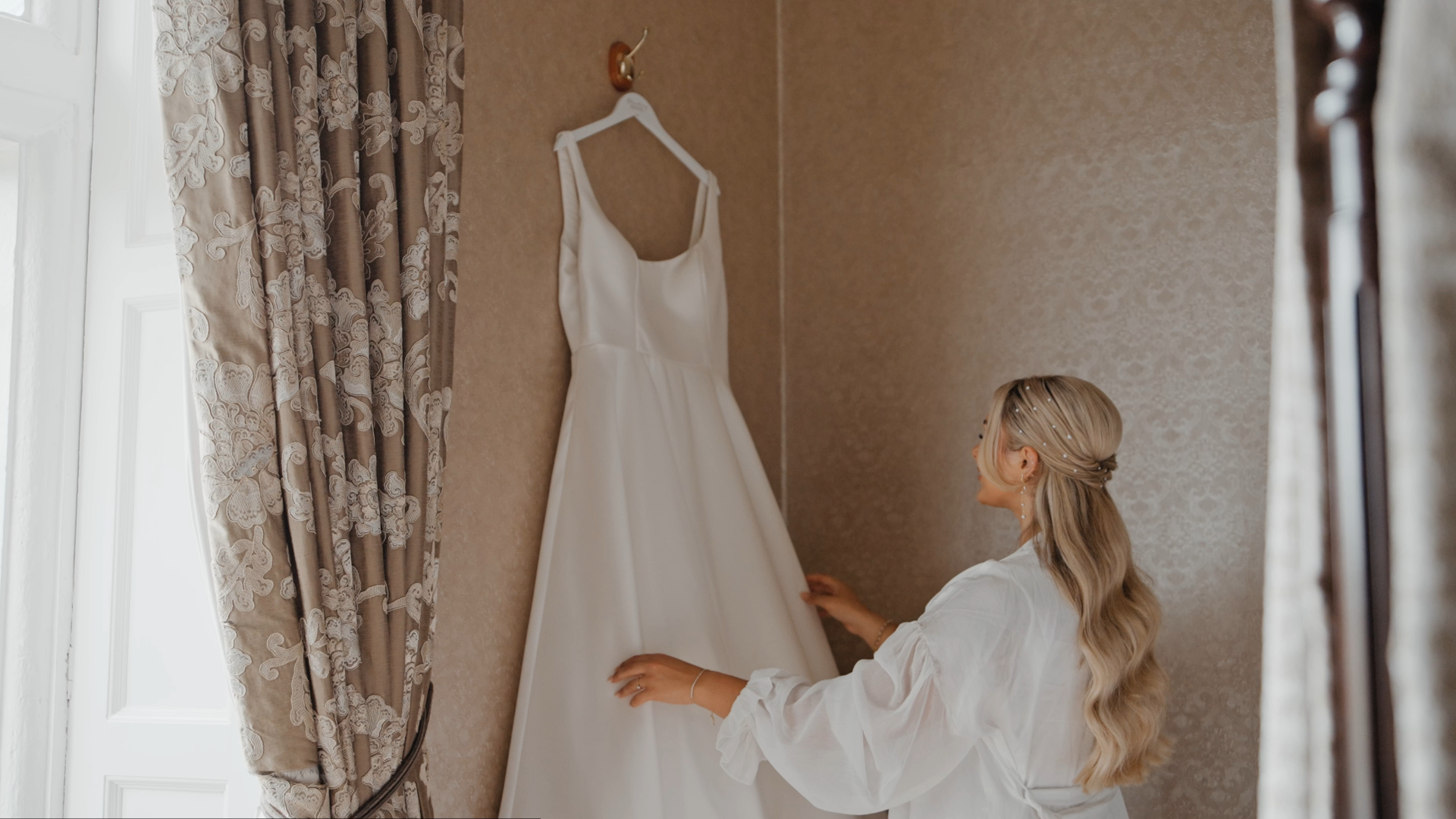 A person with long blonde hair stands in a softly lit room, wearing a white robe. They are reaching towards a white wedding dress hanging on the wall. Patterned curtains frame this serene moment of bridal prep videography.