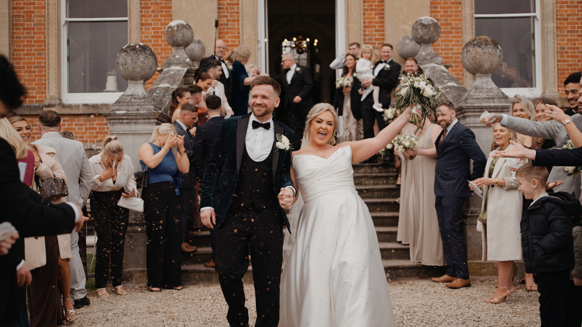 A bride and groom joyfully walk down the steps of Crowcombe Court, surrounded by cheering guests. The bride holds a bouquet, and the groom clasps her hand as confetti rains down. Captured by a talented wedding videographer, everyone is dressed in formal attire for this unforgettable moment.