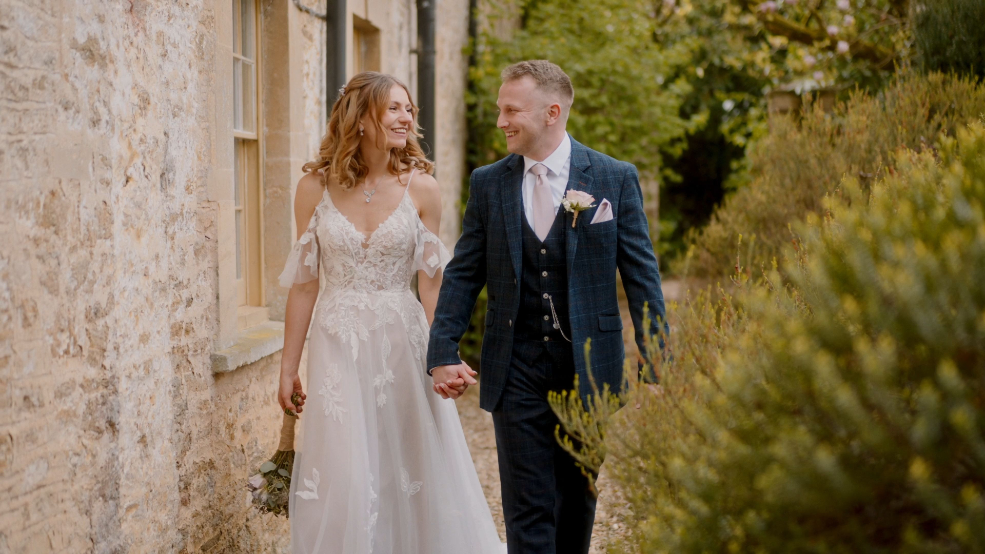 A bride in a white gown and a groom in a dark suit walk hand in hand along the stone pathways of Elmhay Park, surrounded by lush greenery. Their smiles create a joyful, romantic scene perfect for capturing with exquisite wedding videography.