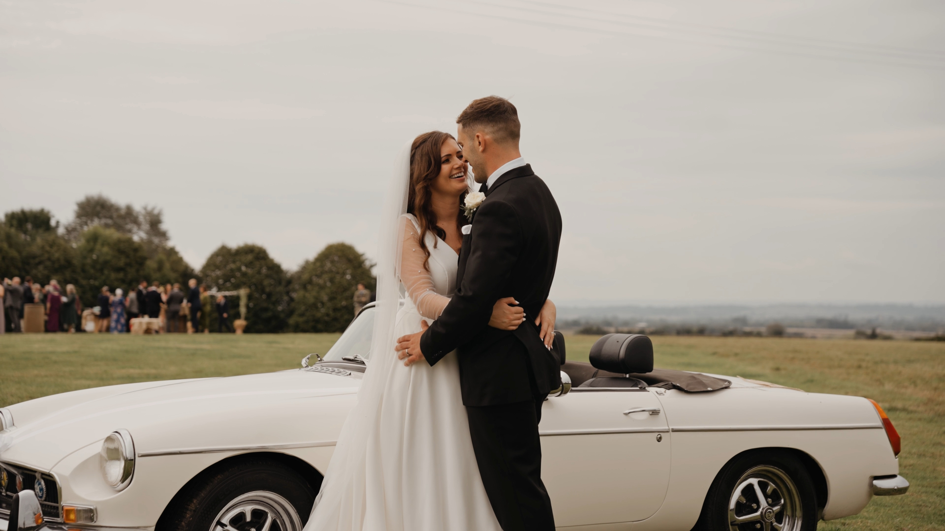 Bride and groom hugging in front of their wedding car, with a scenic green field in the background — captured by a Somerset to Cotswolds wedding videographer.