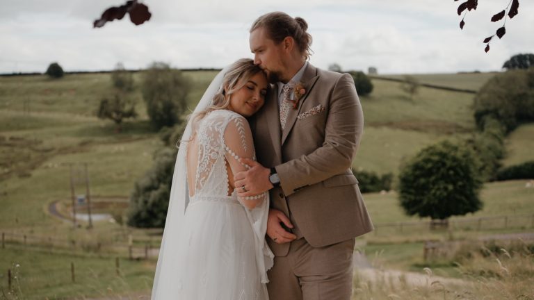 A bride and groom embrace in a grassy, rural landscape. The bride wears a white lace gown, and the groom is in a tan suit. Embracing their waste-free wedding ethos, they stand closely with the groom kissing the brides forehead amidst fields and scattered trees.