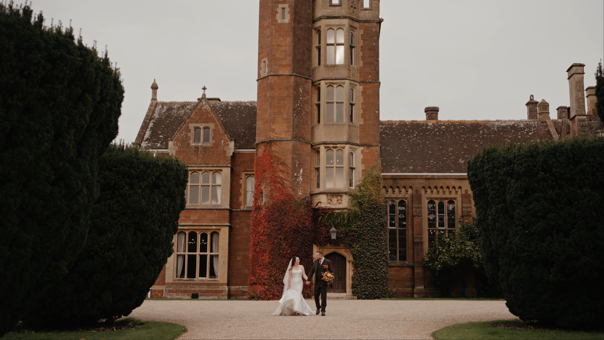 Somerset wedding videographer captures bride and groom walking hand in hand past the iconic red-brick building of St Audries Park.