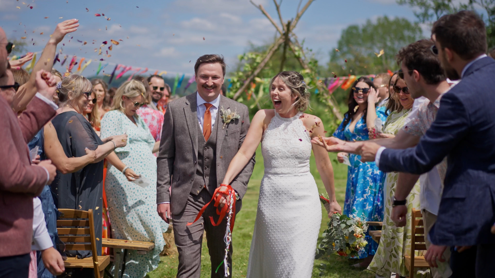 A wedding videographer documents the bride and groom walking hand in hand through a vibrant confetti tunnel at their outdoor wedding, surrounded by cheering guests.