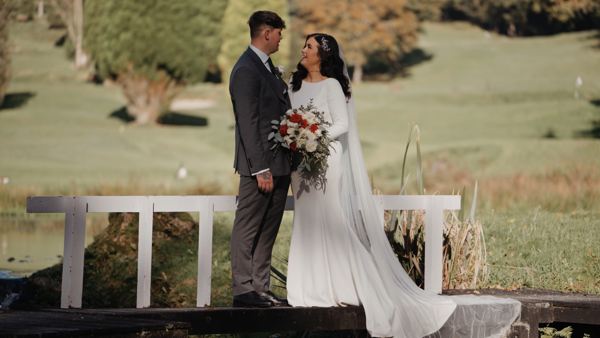 Romantic moment on a wooden bridge as bride and groom smile at each other, surrounded by greenery at a wedding venue in the South West.