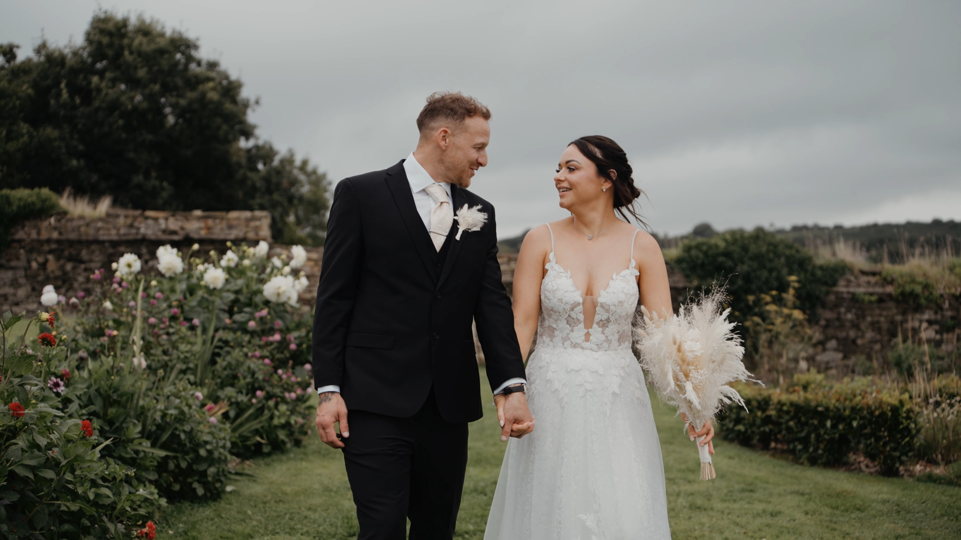 Bride and groom walking through a lush garden with wildflowers and stone walls, a picturesque scene from a South West wedding venue.