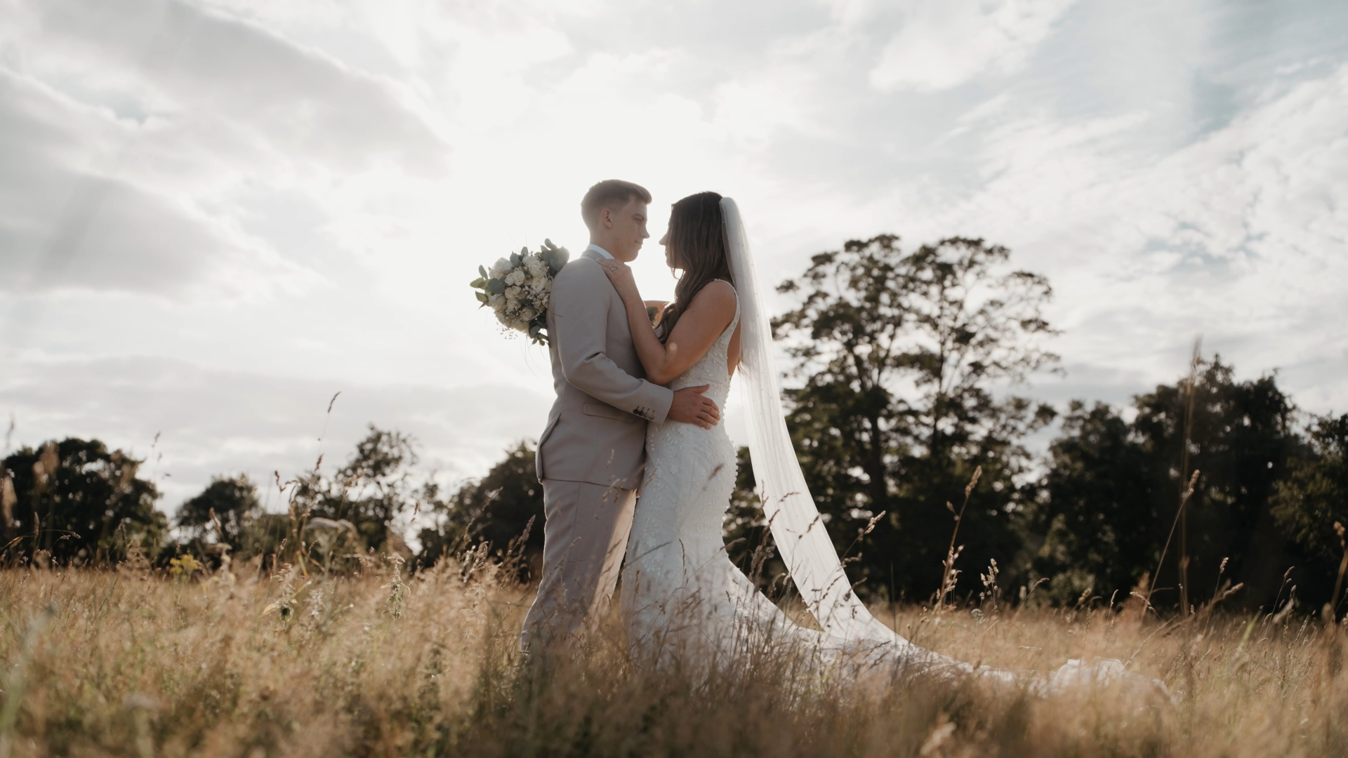 Bride and groom smiling at each other in a grassy field surrounded by tall trees, a natural moment captured by a documentary wedding videographer.