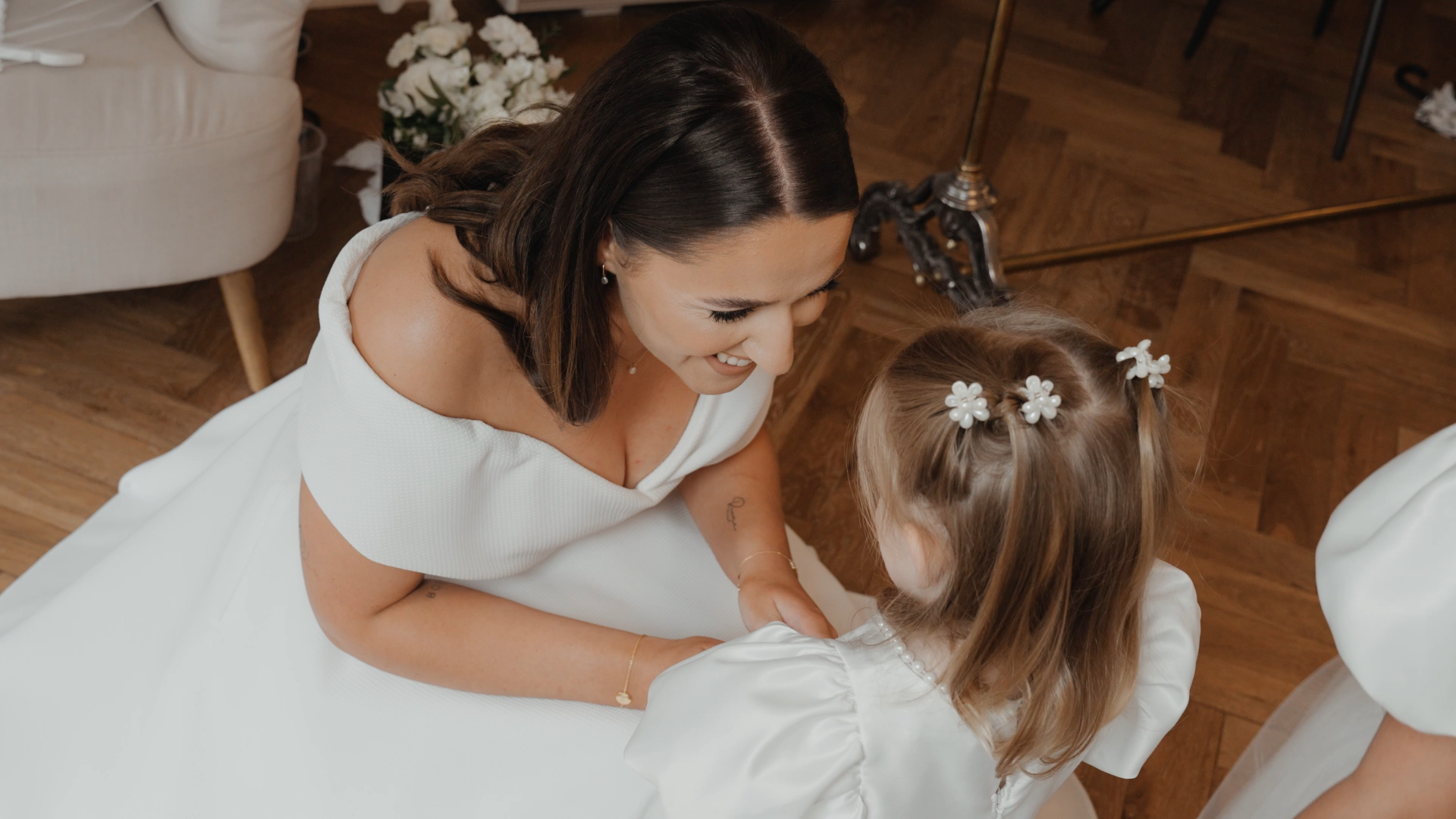 A heartfelt moment captured by a wedding videographer as the bride kneels to hold hands and talk gently with a young girl in soft natural light.