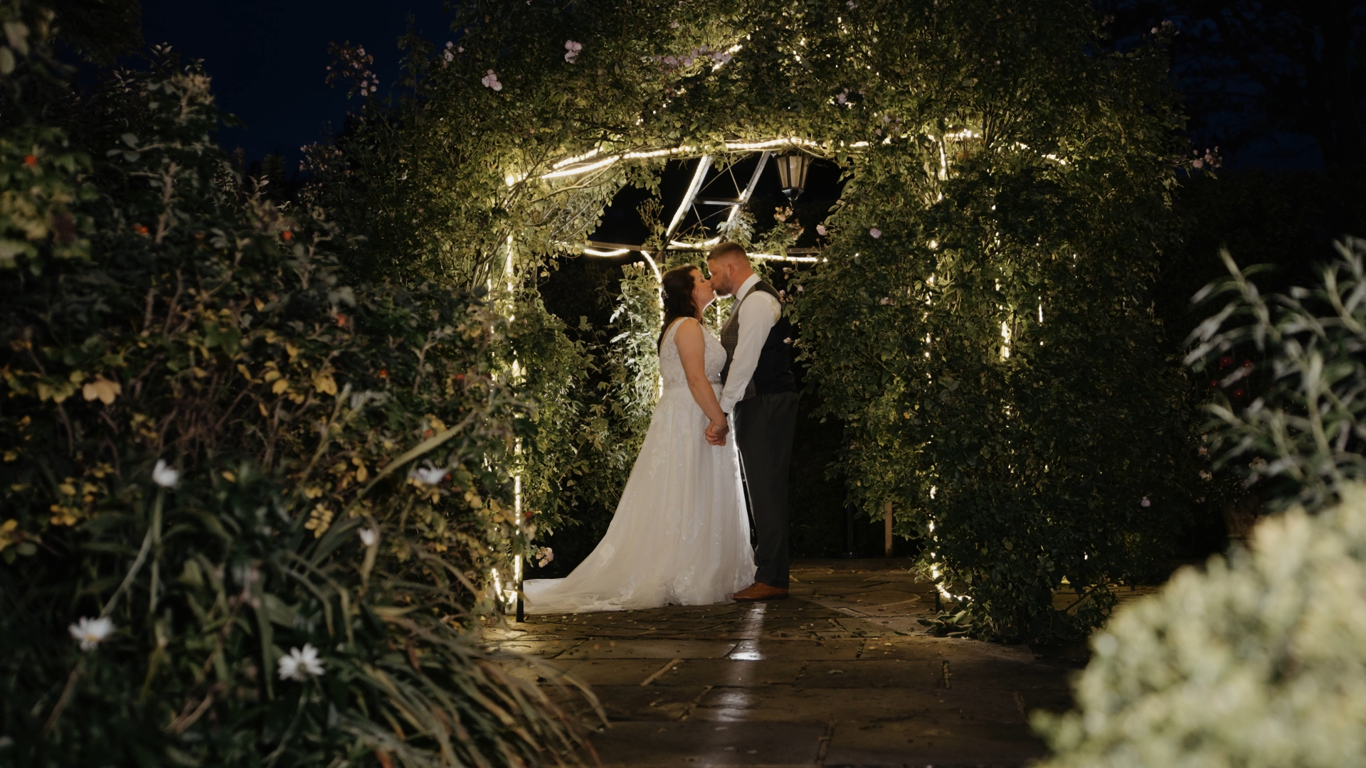 Bride and groom sharing a romantic kiss under a vine-covered shed adorned with twinkling lights, captured at night
