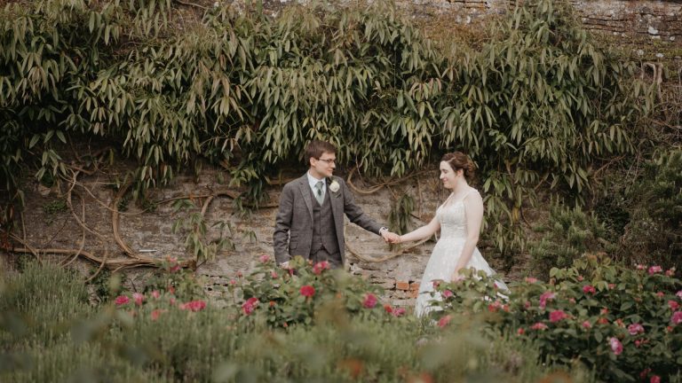 Groom leading the bride by the hand through a garden with a lush leafy background, capturing a romantic moment that reflects natural and intimate wedding trends in 2025.