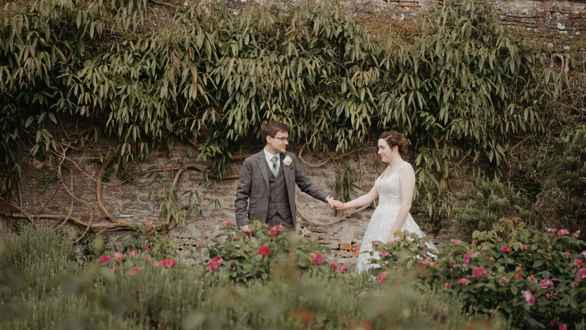 Groom leading the bride by the hand through a garden with a lush leafy background, capturing a romantic moment that reflects natural and intimate wedding trends in 2025.