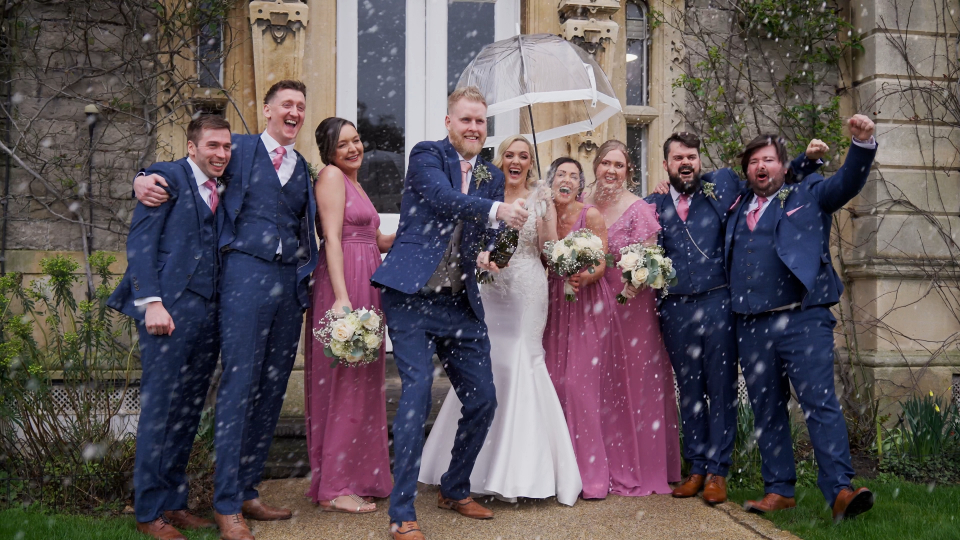 Groom popping champagne as the bride holds an umbrella and the wedding party celebrates in the rain — a lively moment caught by a documentary wedding videographer.