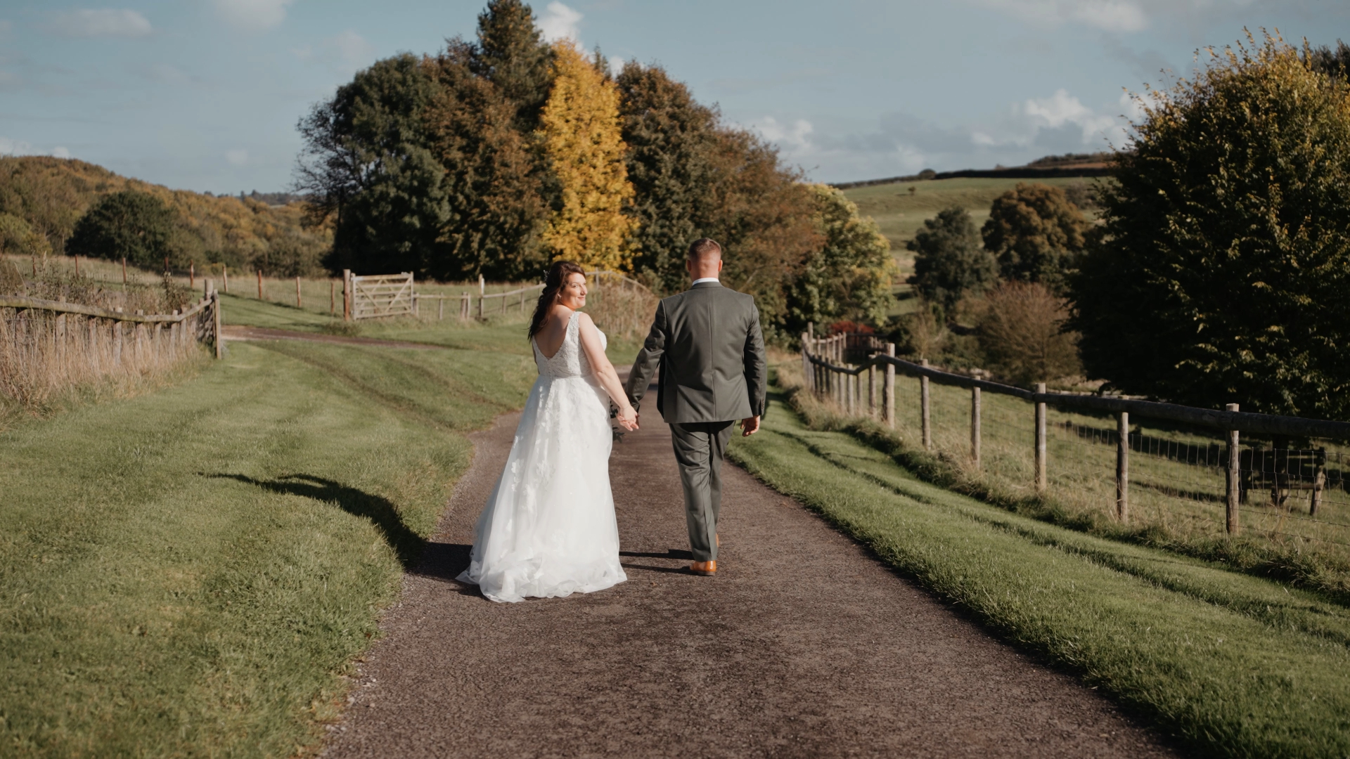 A bride looks back at the camera while holding hands with the groom, walking down a paved path in the middle of a vast field, with a sense of joy and love in the air.