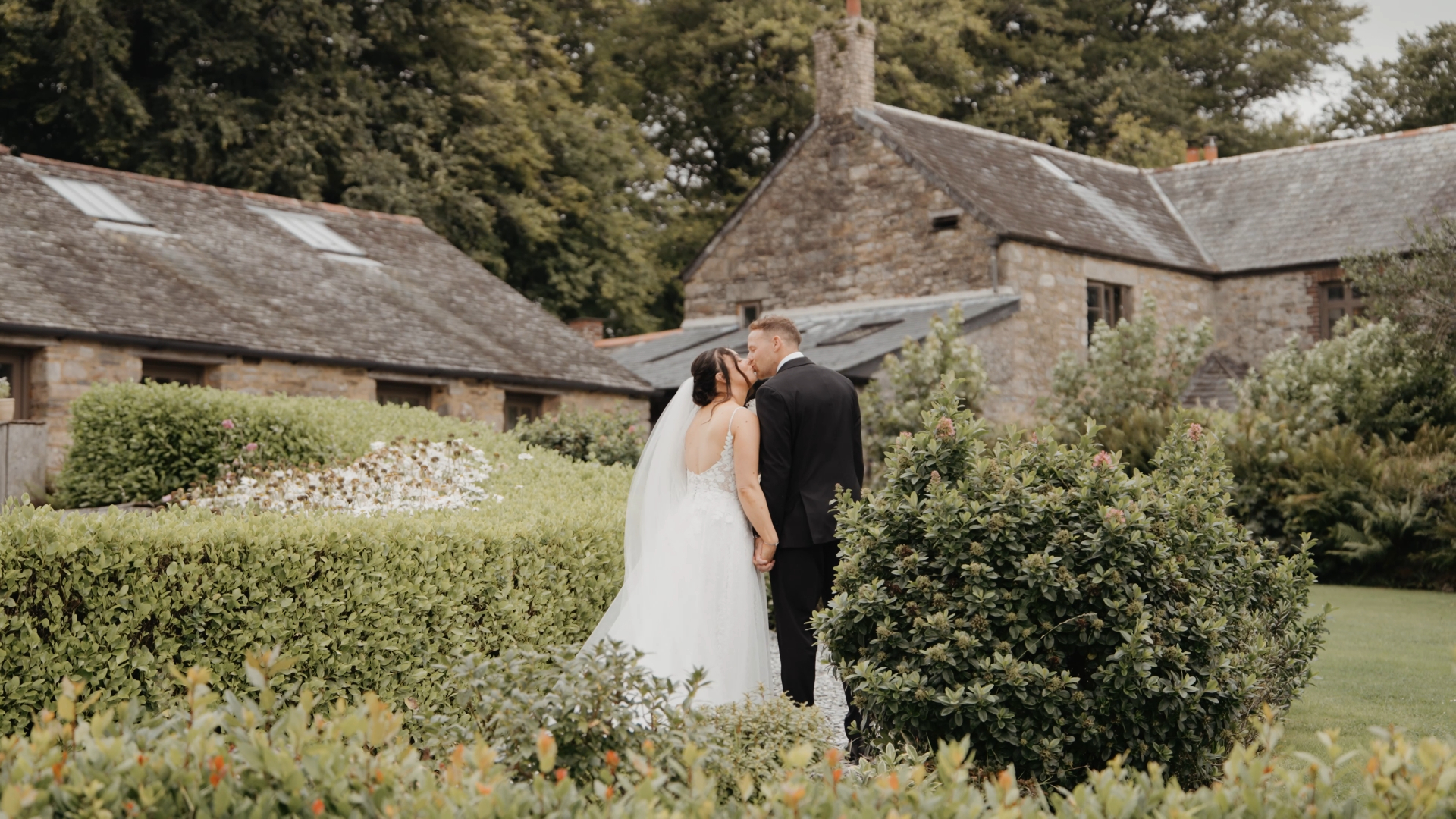 A bride and groom share a tender kiss in a lush garden filled with greenery and blooms — a timeless moment by a Somerset to Cotswolds wedding videographer.