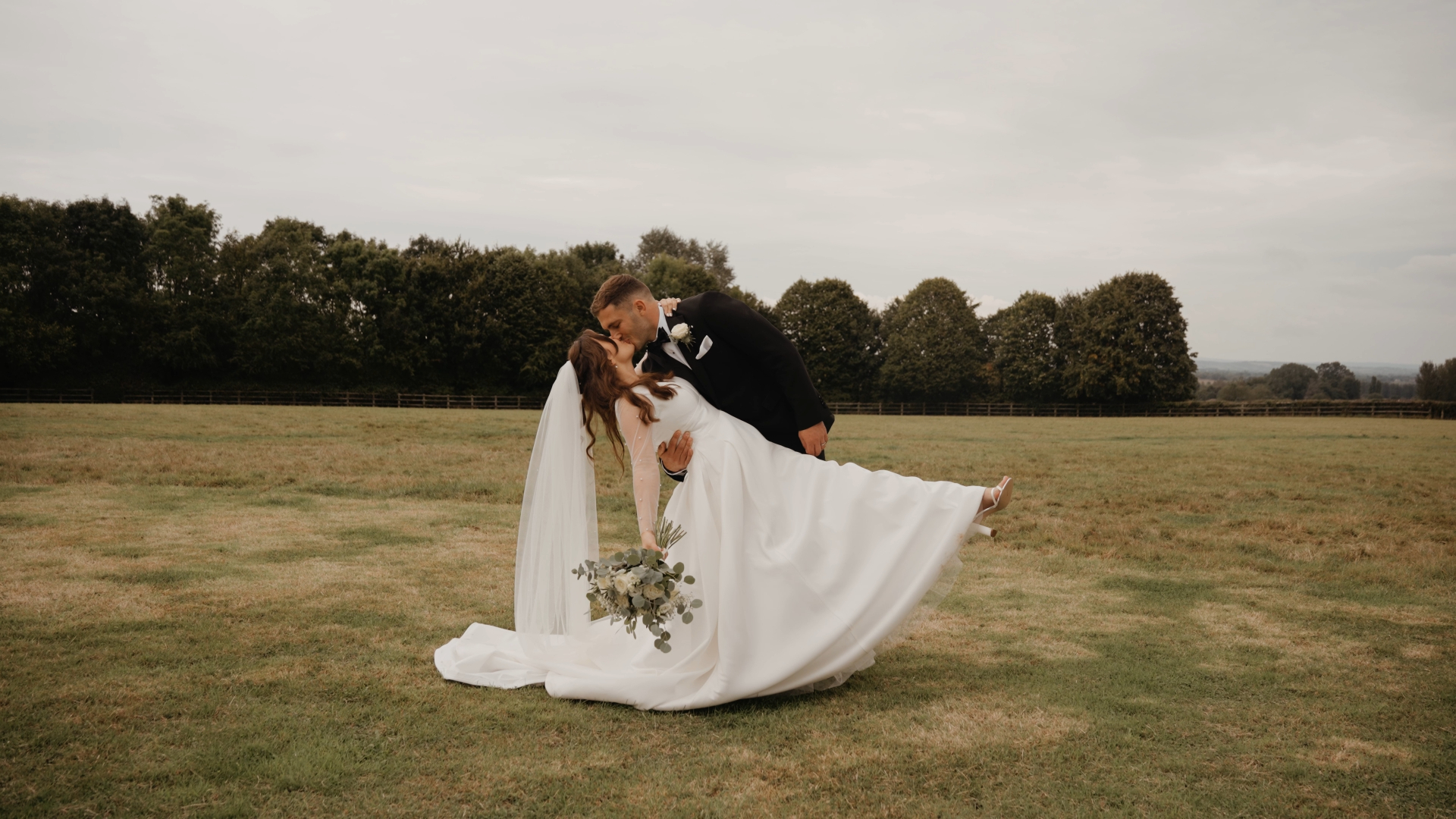 Groom bending the bride gently for a kiss in an open field, surrounded by nature — a romantic shot by a Somerset to Cotswolds wedding videographer.