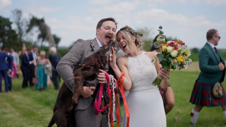 Bride and groom laughing together as groom holds dog during wedding reception.
