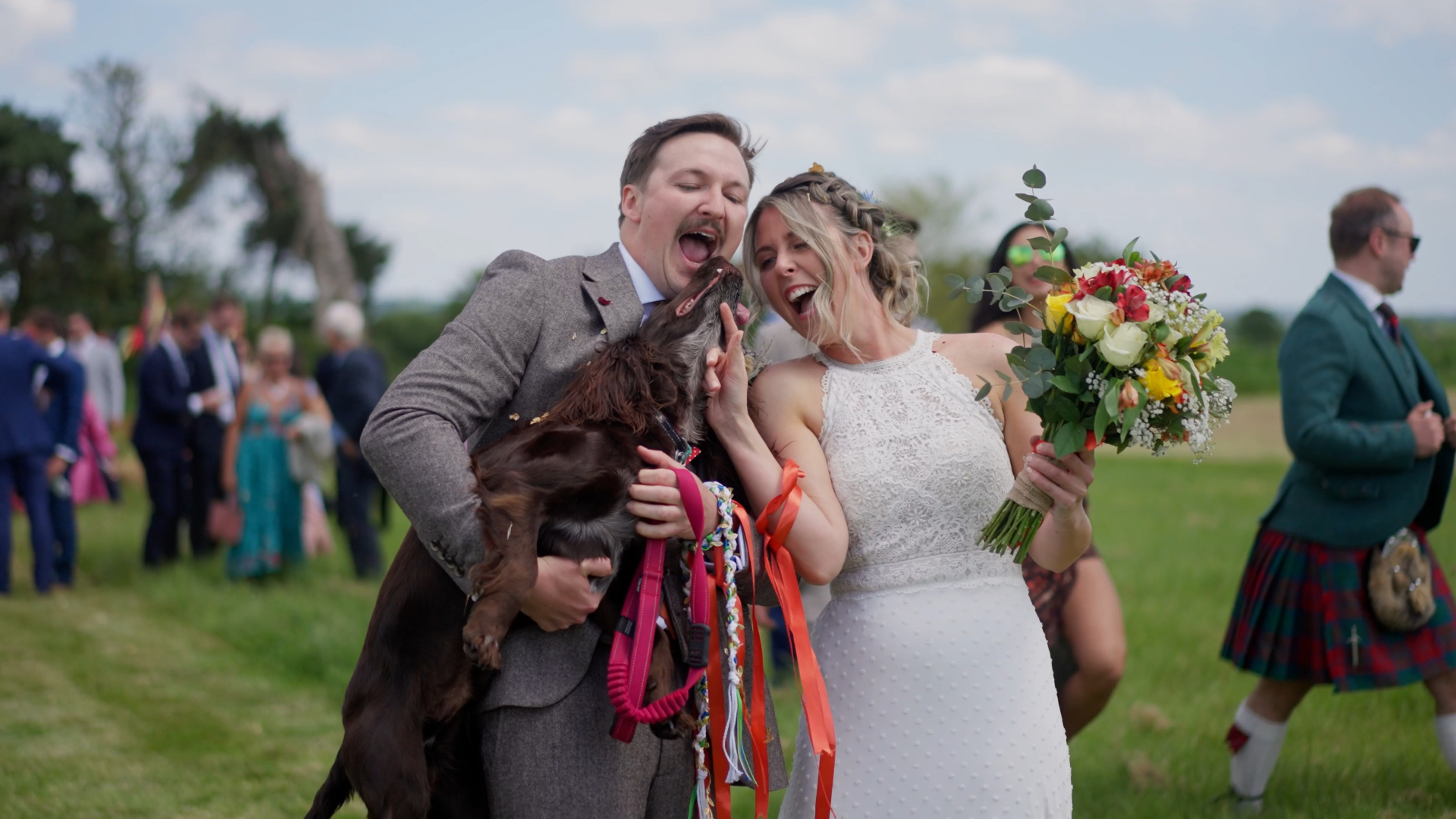 Bride and groom laughing together as groom holds dog during wedding reception.