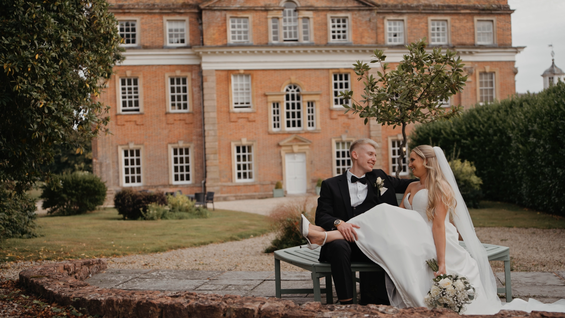 Intimate moment captured by a Somerset wedding videographer as bride and groom relax on a bench outside Crowcombe Court.