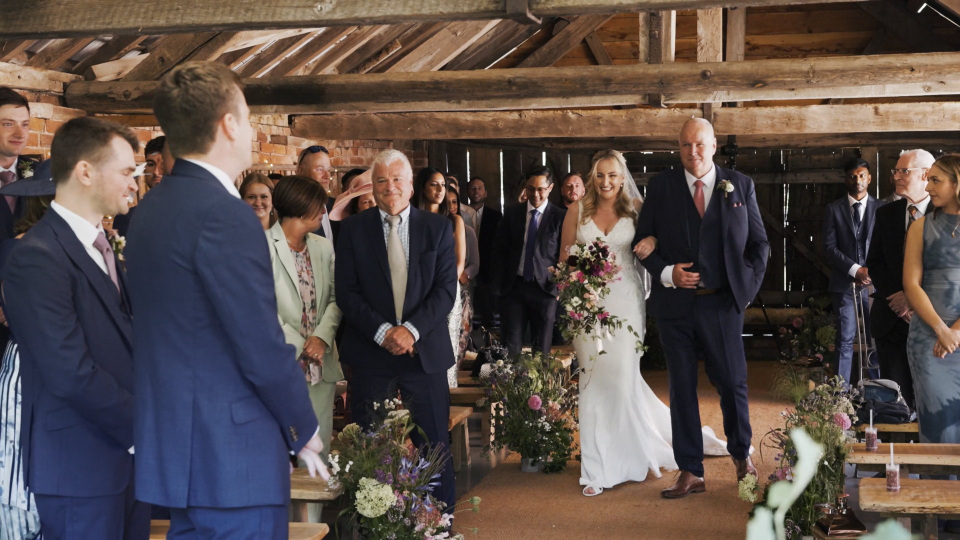 Bride walks down the aisle with her dad at Haybarn as guests look on, captured by a Somerset wedding videographer.