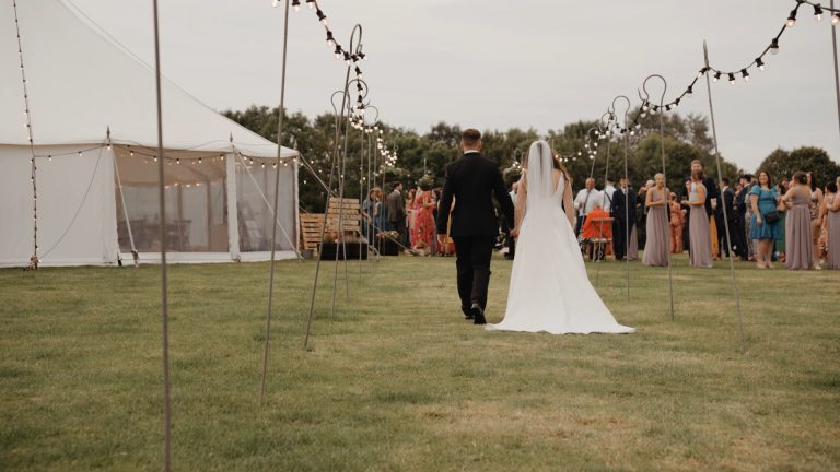 Bride and groom walking hand-in-hand down the middle of a field, with a makeshift aisle lined with glowing fairy lights on either side.