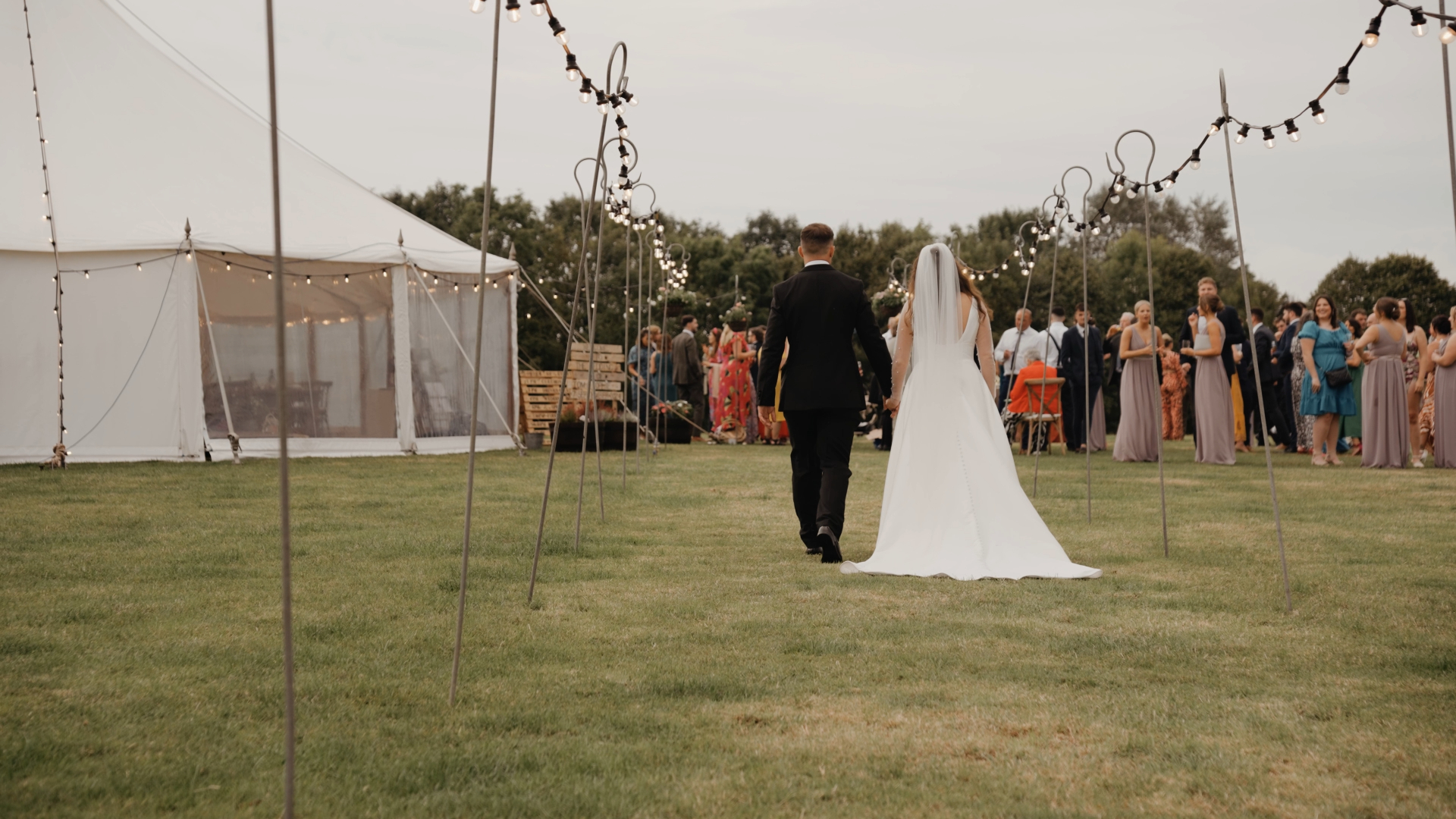Bride and groom walking hand-in-hand down the middle of a field, with a makeshift aisle lined with glowing fairy lights on either side.