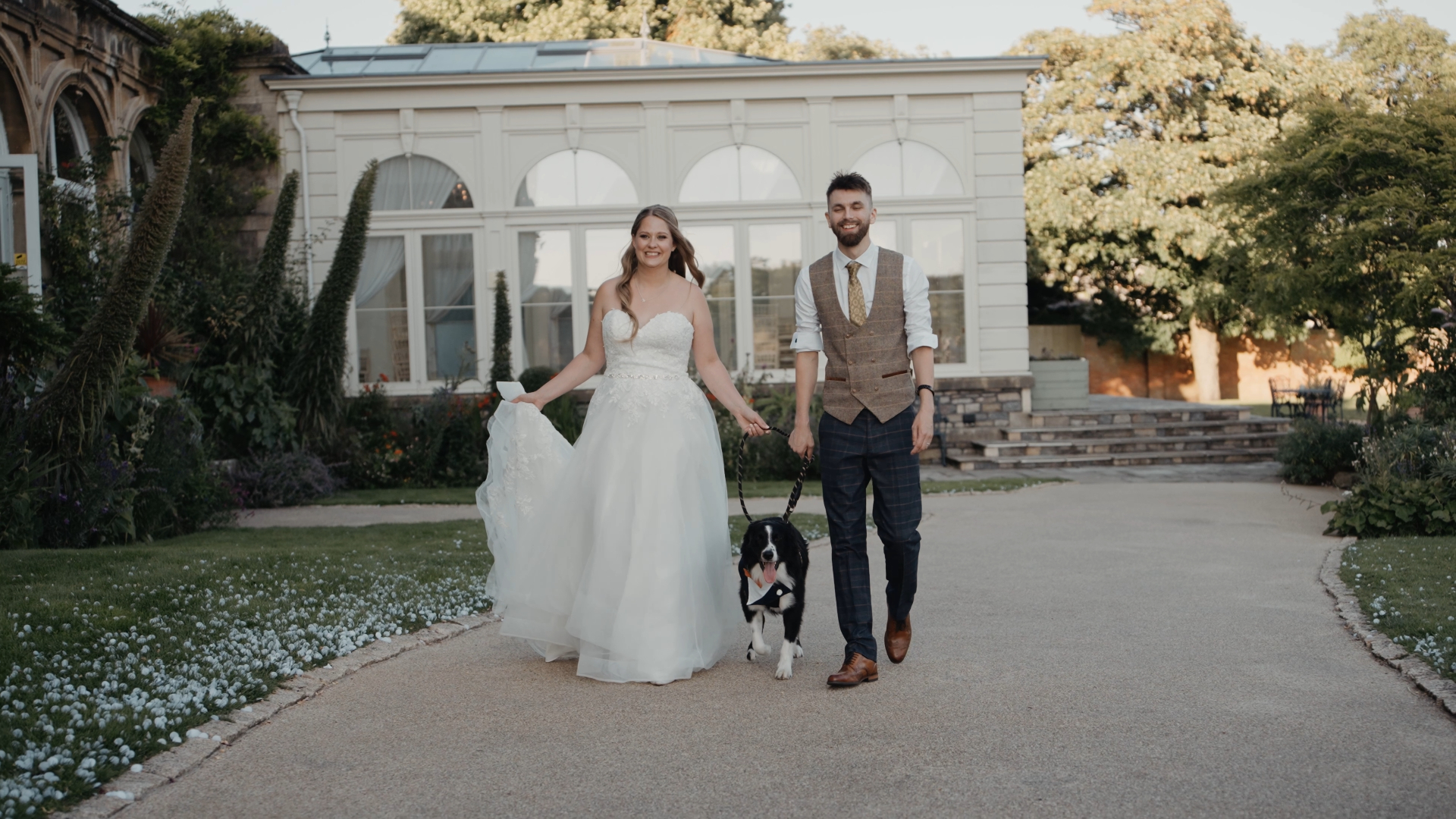 : Bride and groom walking their dog along a pavement, smiling and relaxed, captured beautifully by a Somerset wedding videographer.