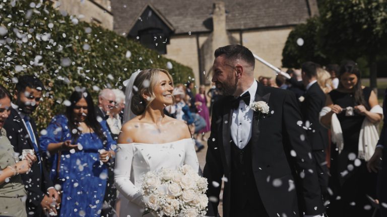 A joyful bride and groom walk hand-in-hand through a flurry of white confetti outside a stone building, surrounded by smiling guests. The bride wears an elegant off-the-shoulder white gown and holds a bouquet of white roses and baby’s breath, while the groom is dressed in a classic black tuxedo with a white boutonnière. Both are looking at each other, smiling brightly, capturing a candid moment of celebration and love.
