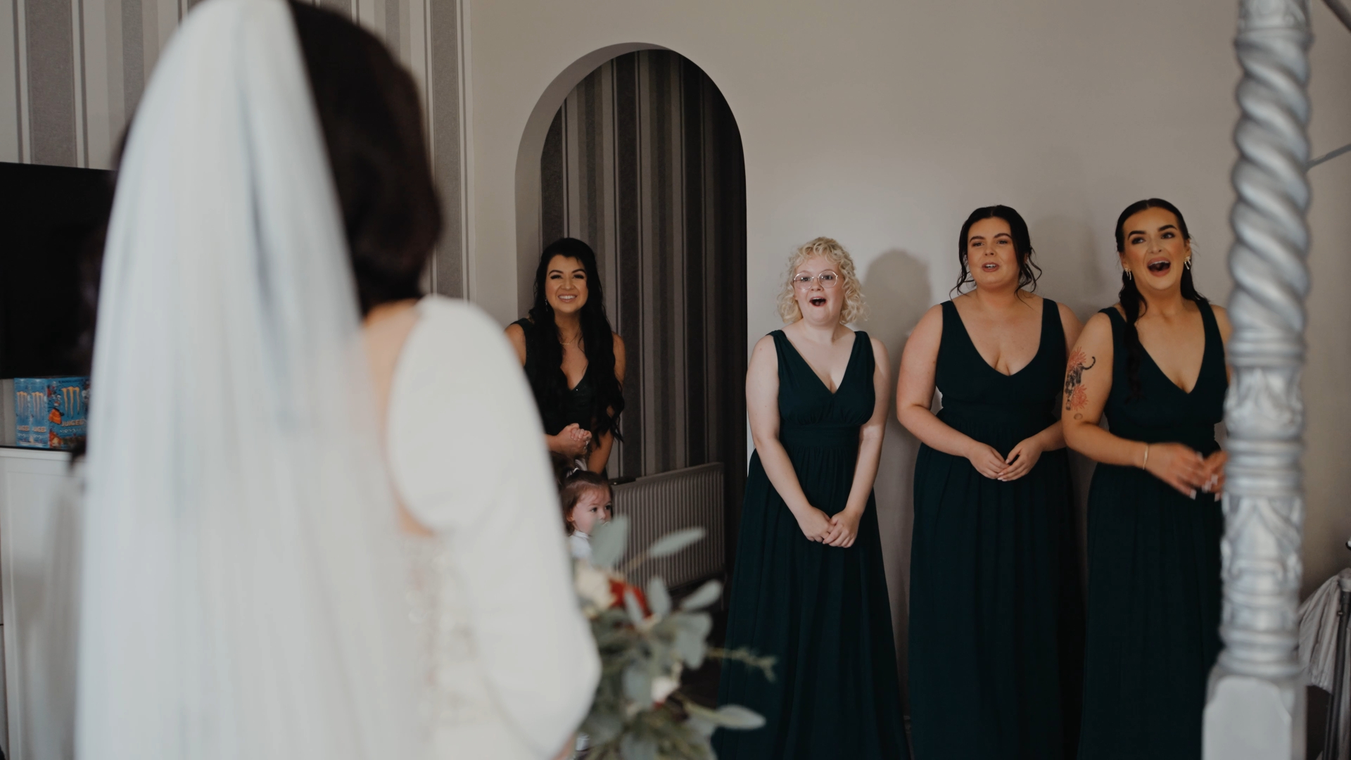A heartfelt first-look moment captured in a documentary-style wedding video as the bride faces her joyful bridesmaids and flower girl in green dresses, surrounded by floral accents and soft light.