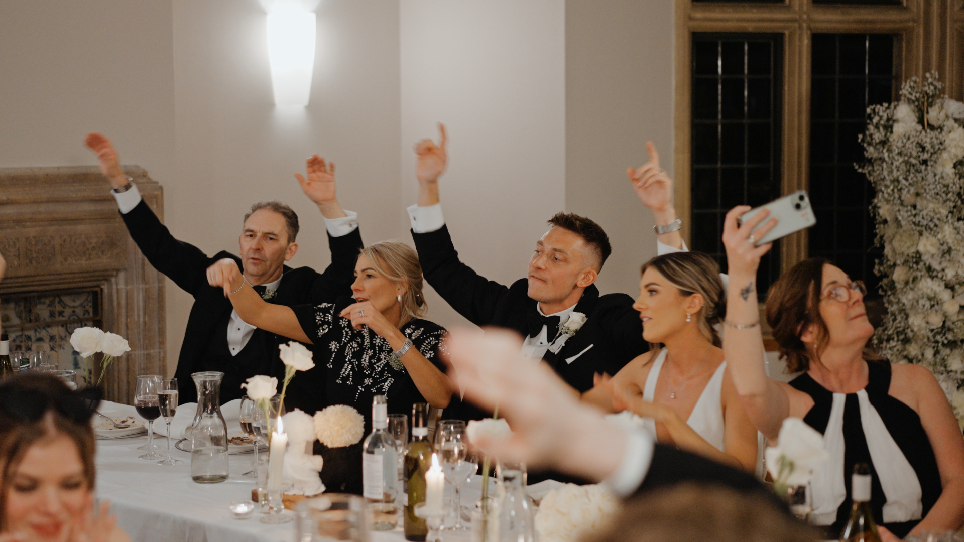 A fun and unscripted moment in a documentary-style wedding video showing the bride and groom dancing while seated, joined by guests raising their hands and laughing in celebration.