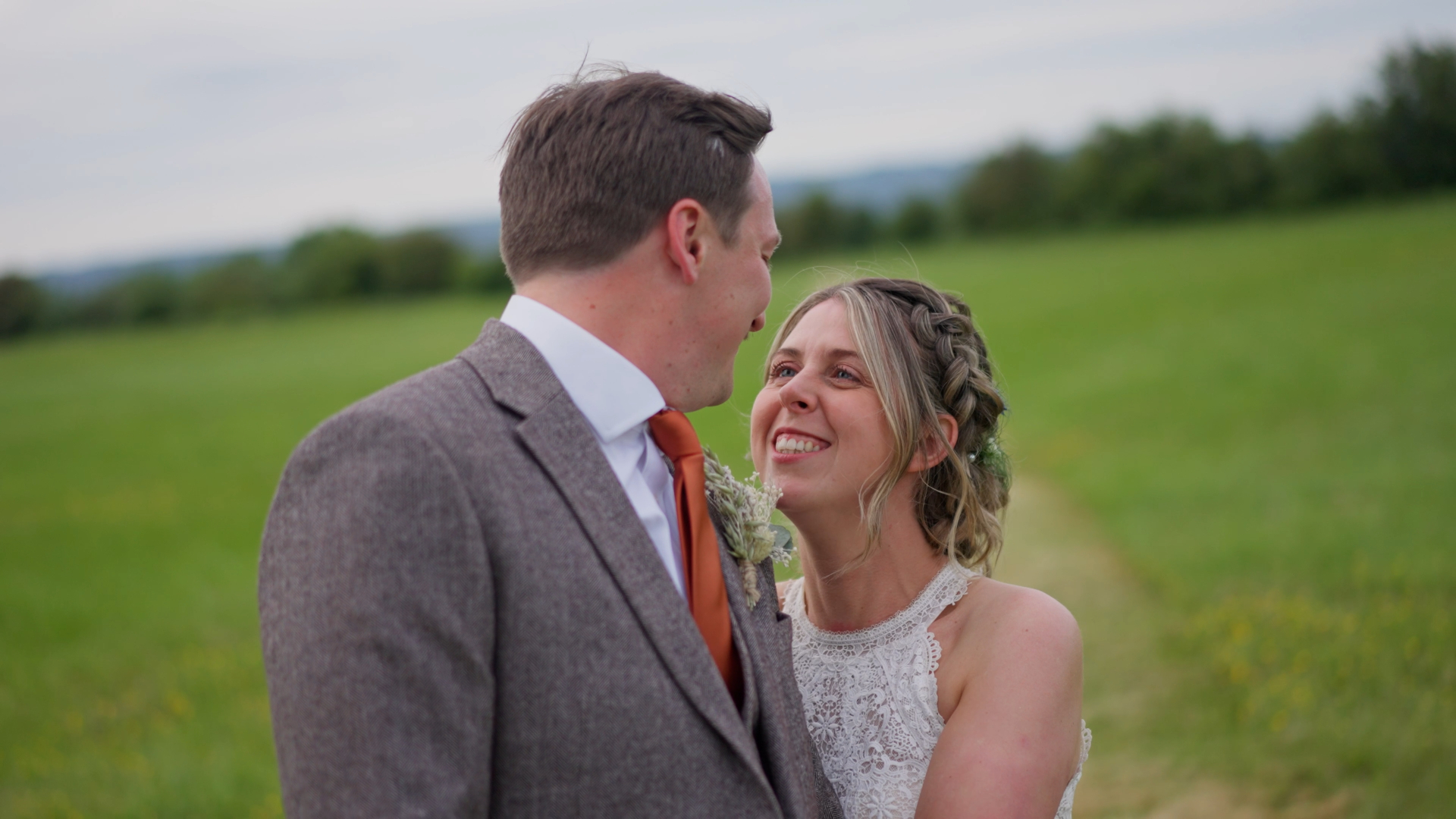 Captured in a documentary-style wedding video, the bride and groom share a genuine smile as they stand in the middle of a vibrant green field under a clear blue sky.