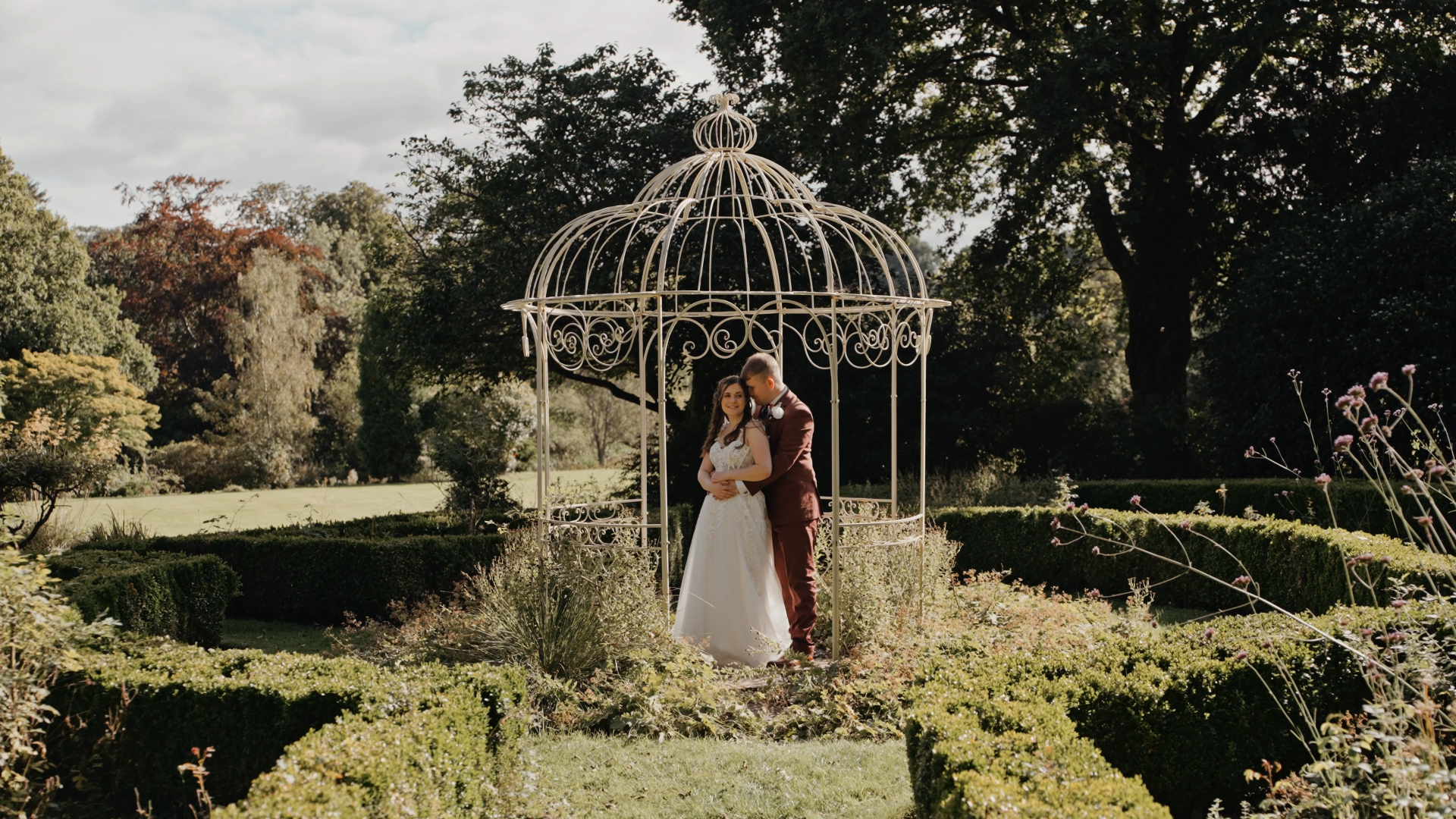 A groom gently backhugging his bride in front of a charming white gazebo in a blooming garden in Somerset, capturing a tender elopement moment surrounded by flowers and greenery.