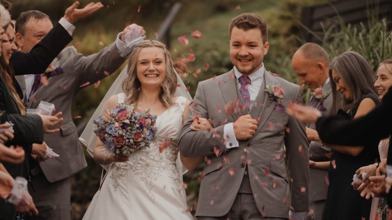 A smiling bride and groom walk arm-in-arm outdoors, celebrating their perfect wedding planning as guests throw flower petals. The bride holds a bouquet in her white dress; the groom sports a light gray suit with a purple tie.