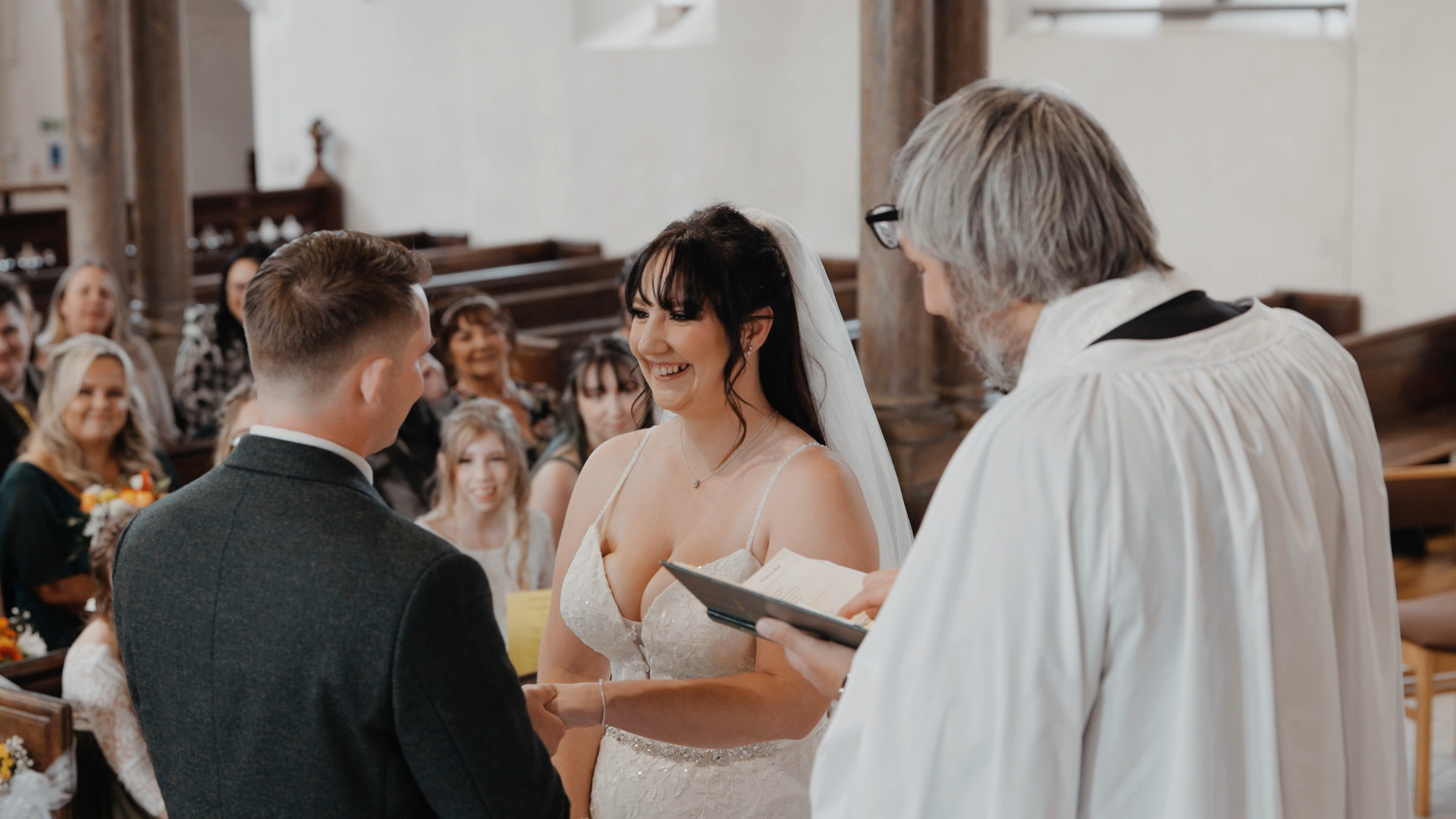 A bride and groom stand facing each other, smiling, as they hold hands during a church ceremony. As the priest reads from a book, they prepare to share emotive wedding vows while guests watch joyfully from the pews.
