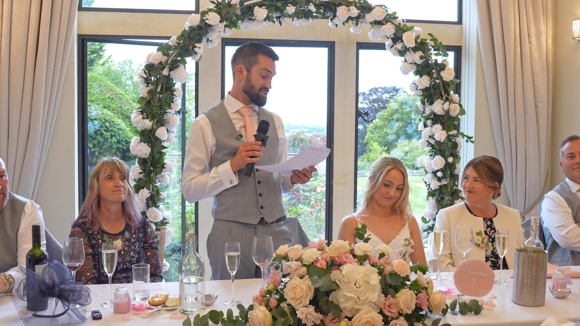 The groom reading his heartfelt speech to the bride during a wedding reception, standing with a microphone as guests look on, capturing an emotive wedding vows moment filled with love and joy.