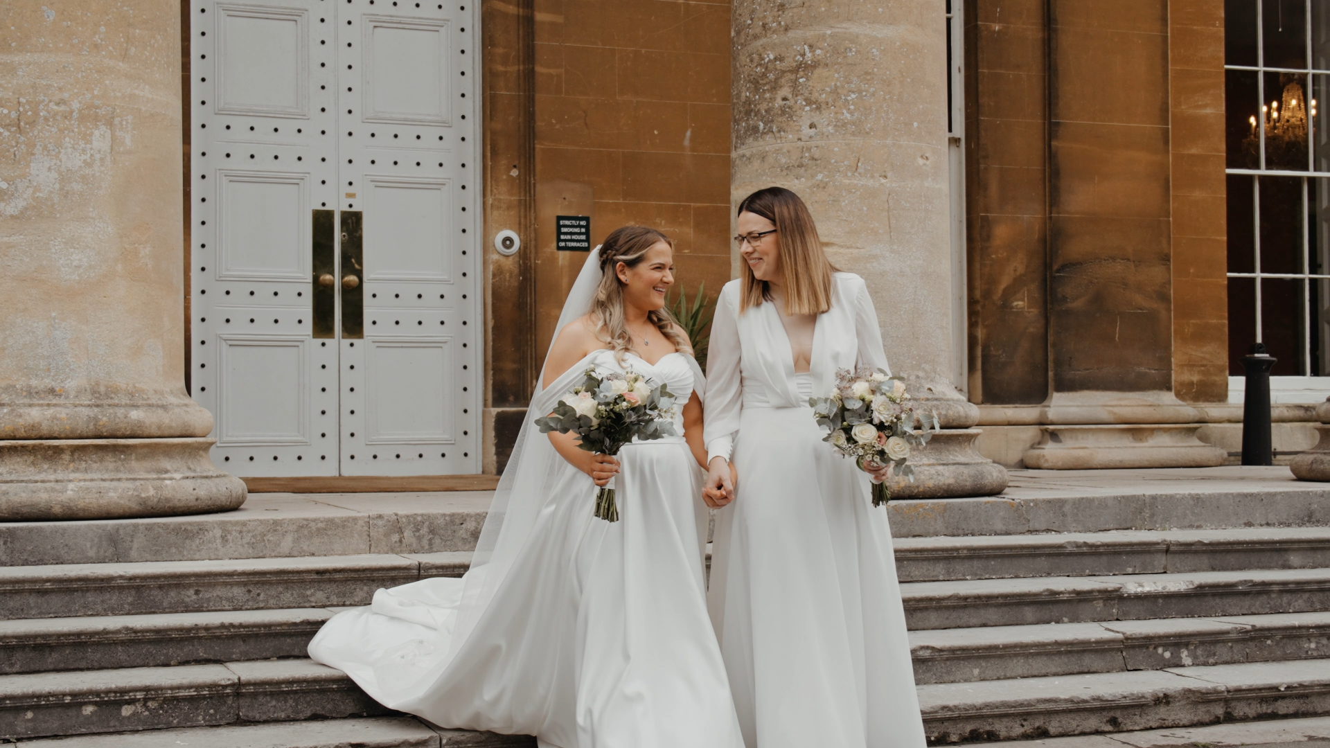 Two brides smiling joyfully as they exit the wedding venue hand in hand, a celebratory moment beautifully captured for their wedding video.