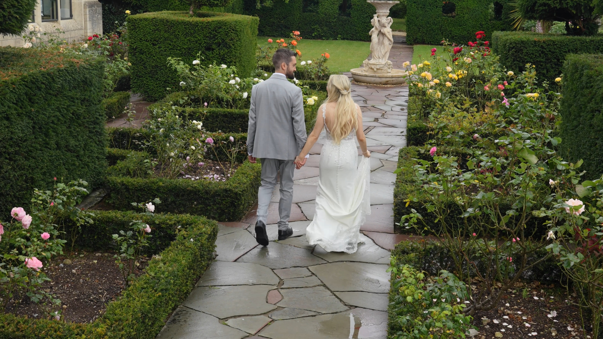 The bride and groom walk hand in hand through a lush garden, sharing a tender moment captured by the wedding videographer.

