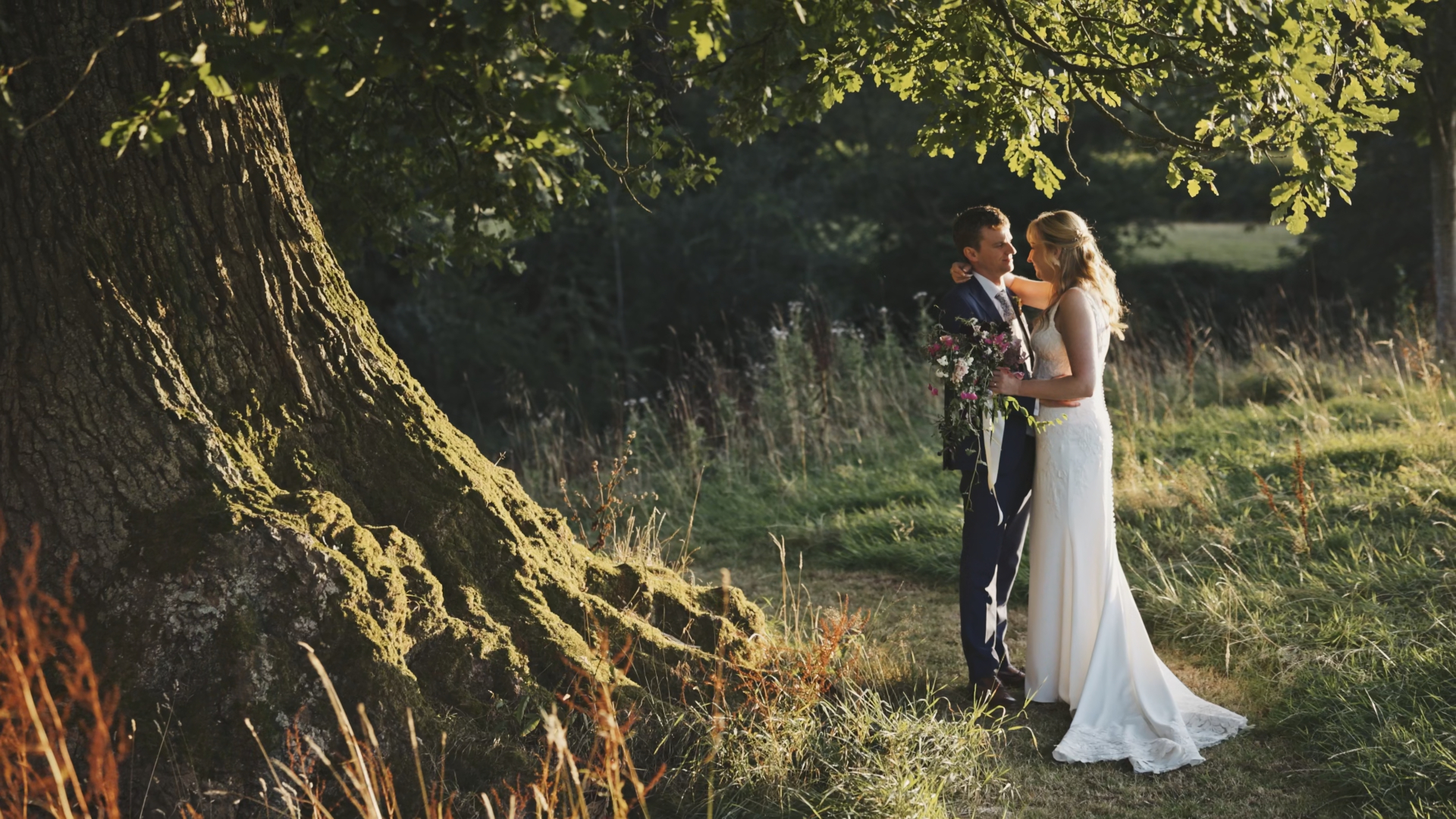  A bride and groom slow dancing beside a large tree in a serene forest setting in Somerset, sharing a quiet, romantic moment during their elopement with soft sunlight filtering through the branches.