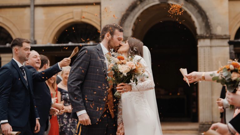 The bride and groom share a kiss as red and orange petals are thrown around them, with the bride holding a vibrant summer-colored bouquet—perfectly capturing the essence of a seasonal wedding celebration.