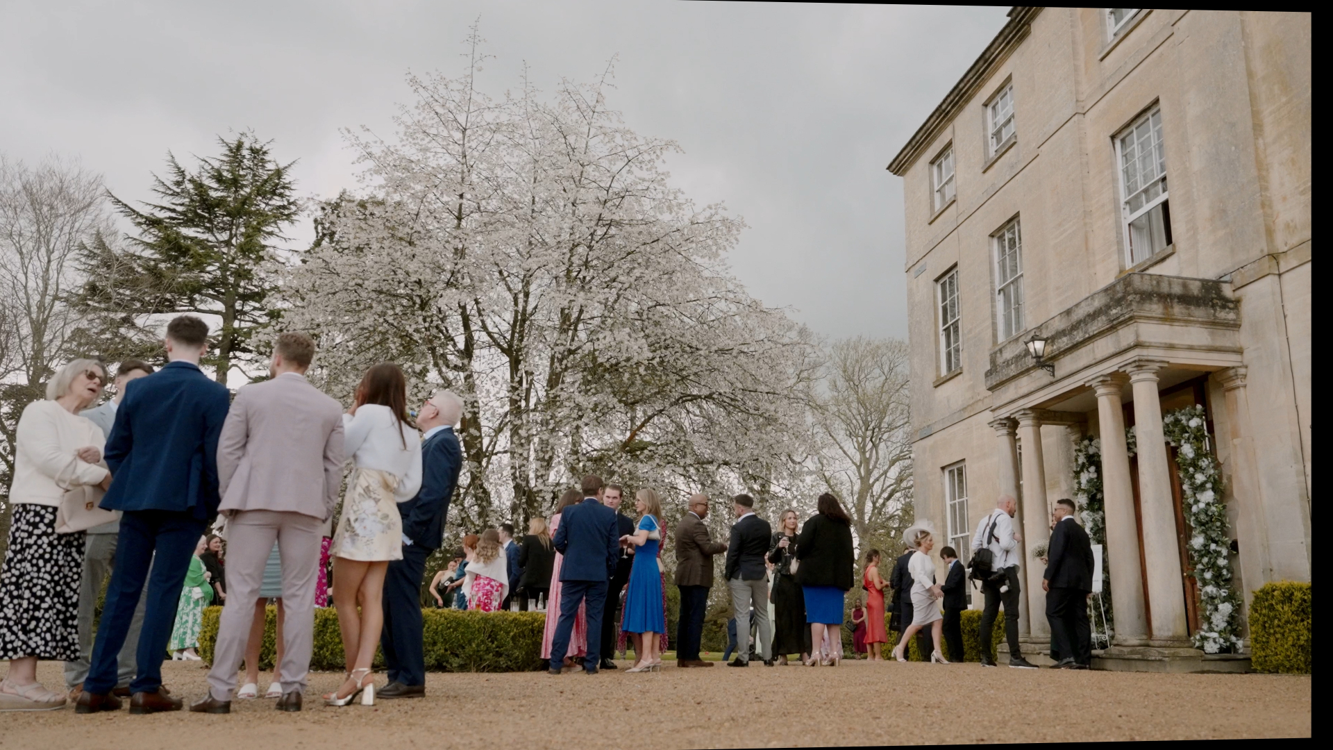 
Wedding guests chatting outside the venue beneath trees covered in white blossoms, capturing a light and airy atmosphere that reflects the charm of a seasonal wedding.
