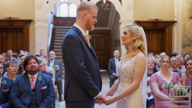 A bride and groom stand facing each other, holding hands as they exchange emotive wedding vows during their ceremony in a grand hall, with guests seated and watching attentively in the background.