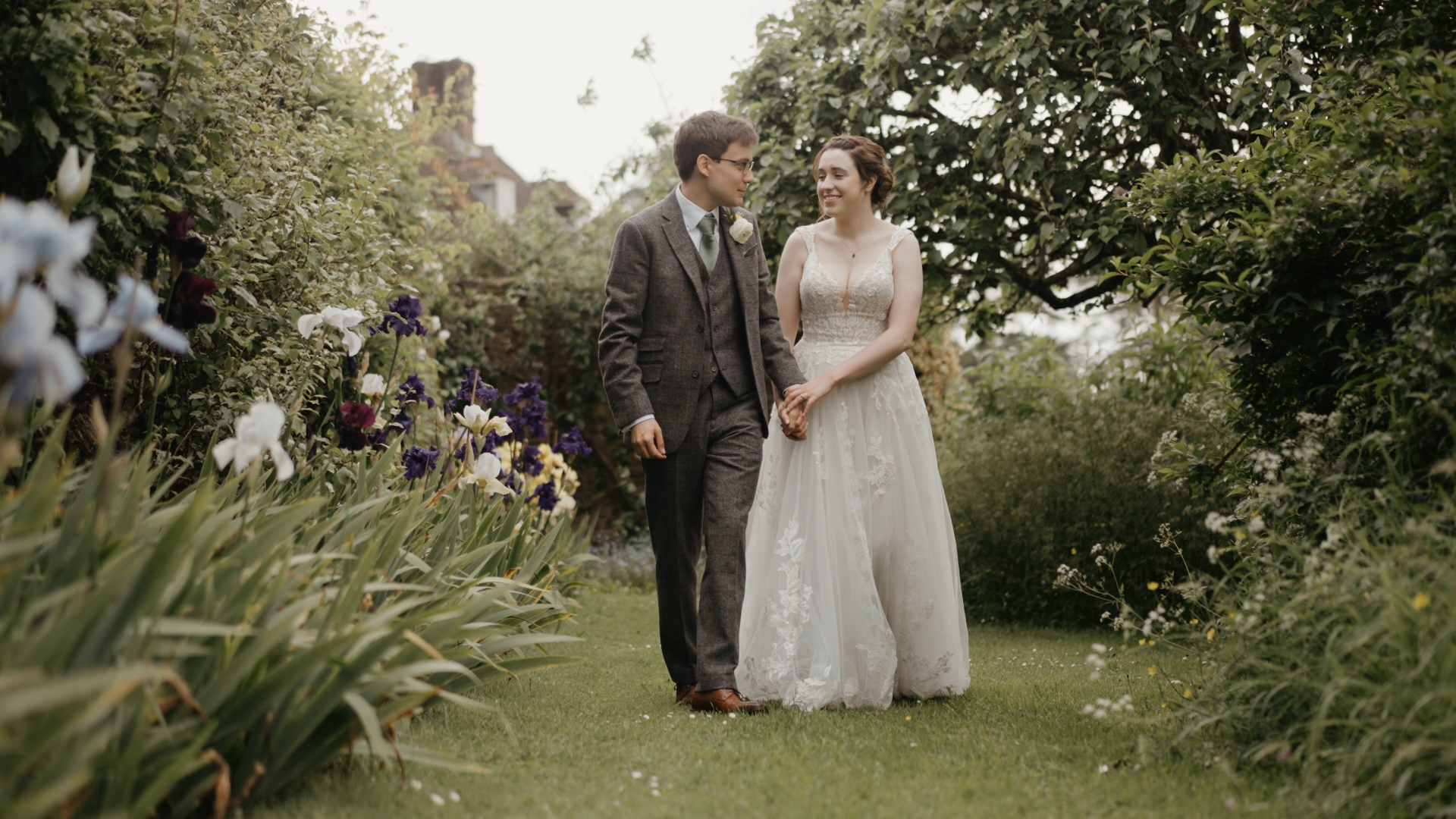 The bride and groom walk hand in hand through a lush garden filled with blooming blue and white flowers, a serene and romantic scene that highlights the beauty of a seasonal wedding.