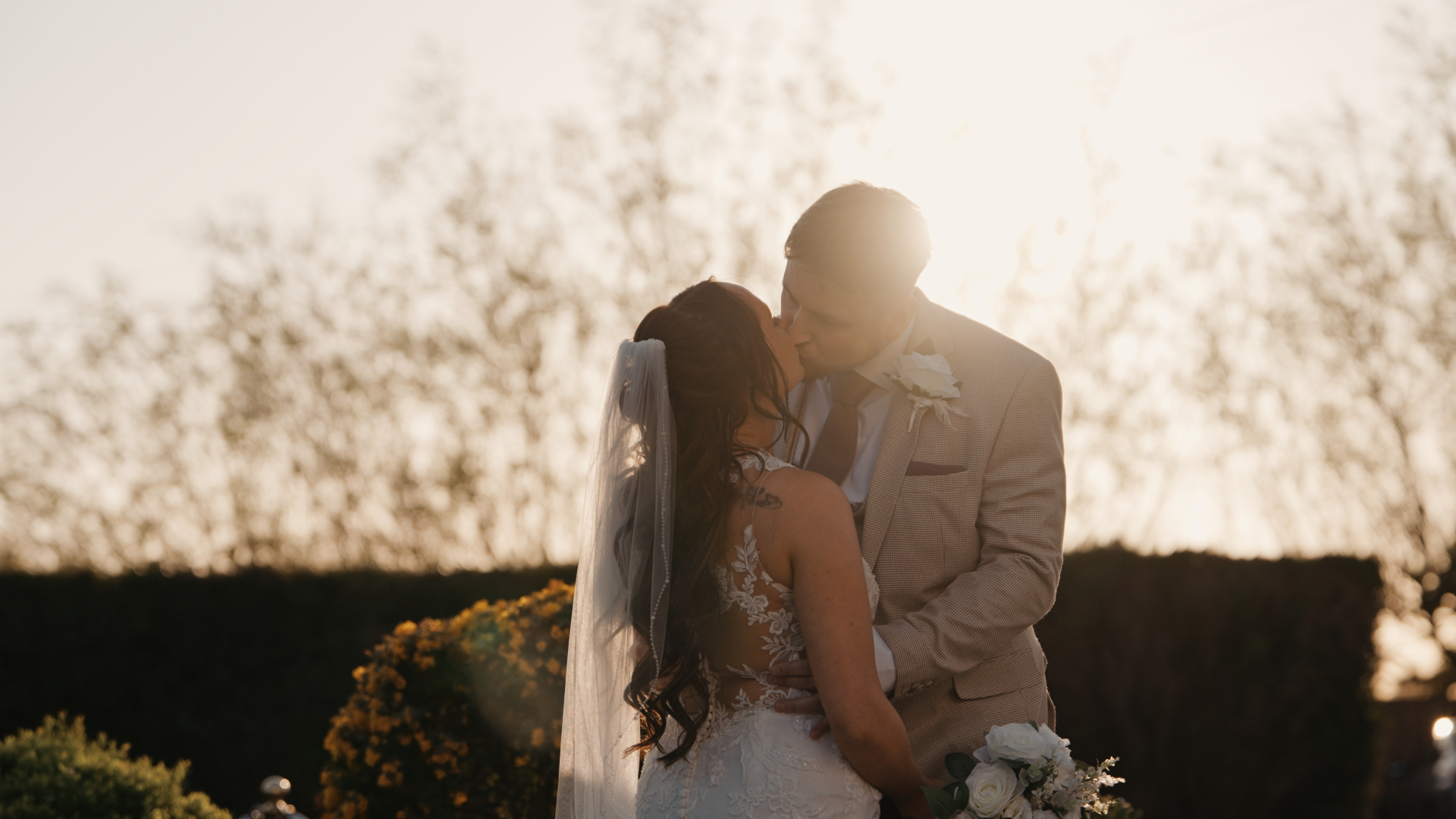 The groom kisses the bride as the sun sets behind them, casting a golden glow over the scene—a romantic moment that can be affected by common wedding videography mistakes like incorrect exposure or missed timing.