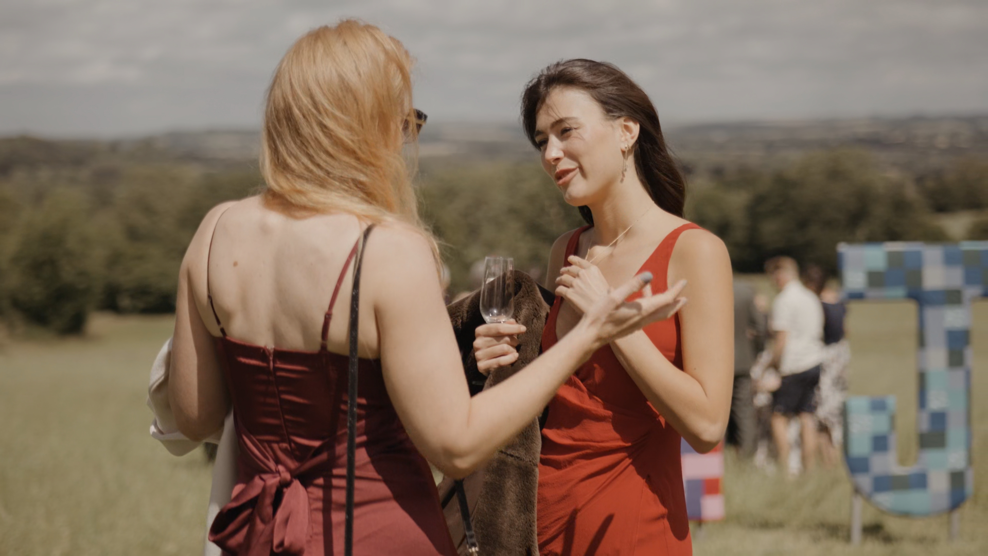 Two female wedding guests wearing burnt orange sleeveless dresses, standing outdoors and enjoying the celebration, their attire perfectly complementing the warm tones of a seasonal wedding.