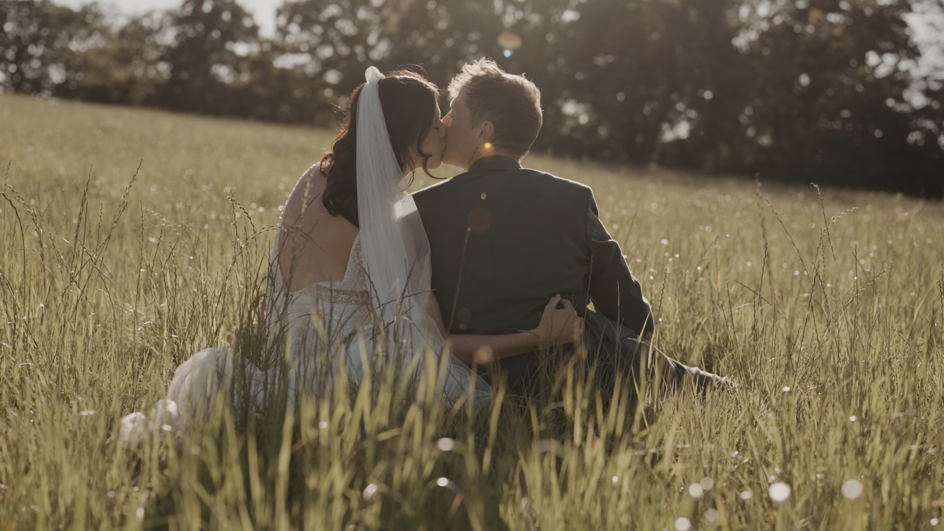 A bride and groom sharing a kiss while sitting in an open field in Somerset, bathed in warm sunlight, capturing a golden, romantic moment during their elopement surrounded by tall grass and nature.