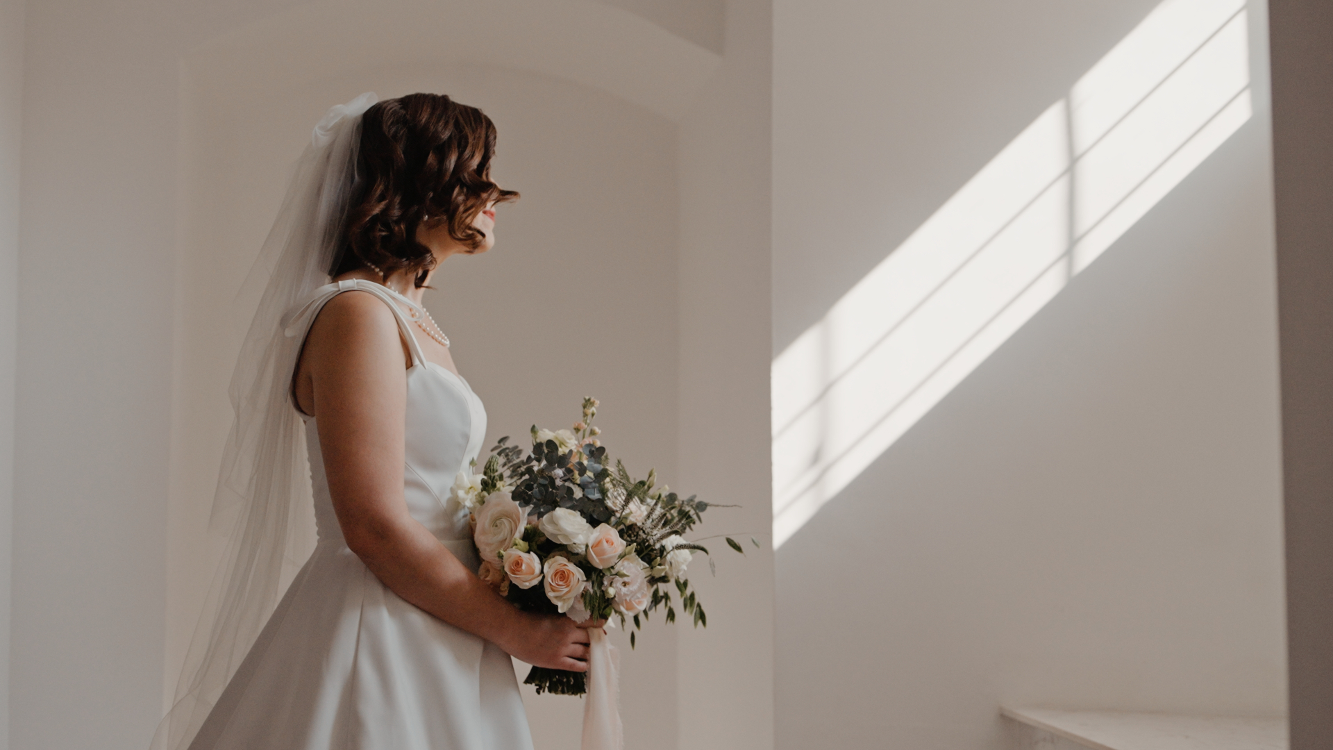 The bride in her wedding dress, holding a bouquet of flowers, gazes thoughtfully out of a window, a candid moment beautifully captured by the wedding videographer.